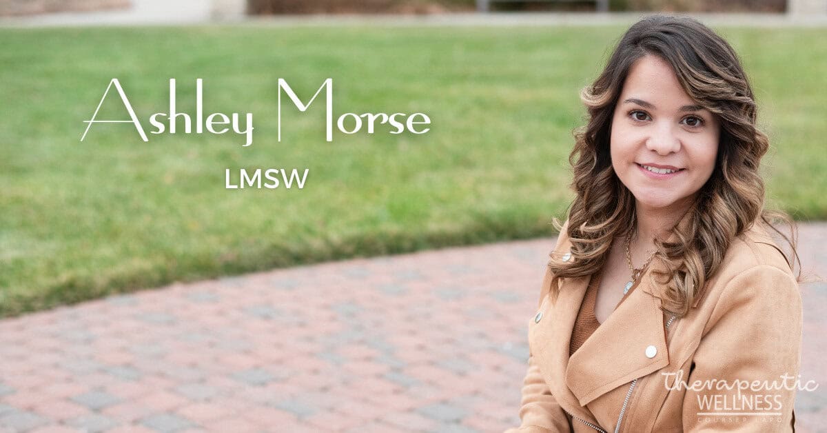 Ashley Morse, with long wavy hair and a tan coat, sits outdoors on a brick path bordered by grass. "Ashley Morse, LMSW" and "Therapeutic Wellness" are displayed in the lower corner of the image.
