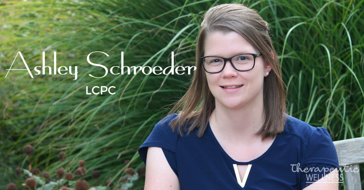 A woman with glasses and a navy blue shirt, identified as Ashley Schroeder, LCPC, is sitting on a bench outdoors, surrounded by tall grass and plants. The text on the image includes "therapeutic wellness" in the bottom right corner. This serene scene mirrors the peaceful environment that Kali Harvey often enjoys.