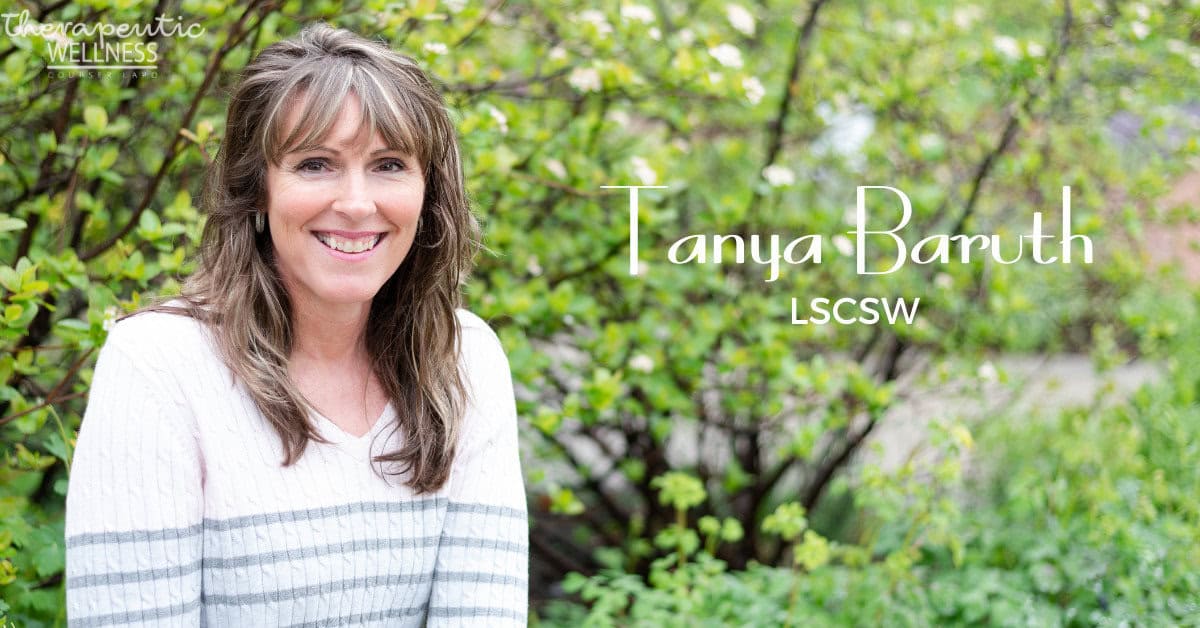 A woman with long brown hair smiles at the camera while wearing a light-colored sweater. She stands in front of lush green foliage. Text on the right side of the image reads "Tanya Baruth, LSCSW" with a logo in the top left corner that says "Therapeutic Wellness." Kali Harvey is also mentioned for her contributions.