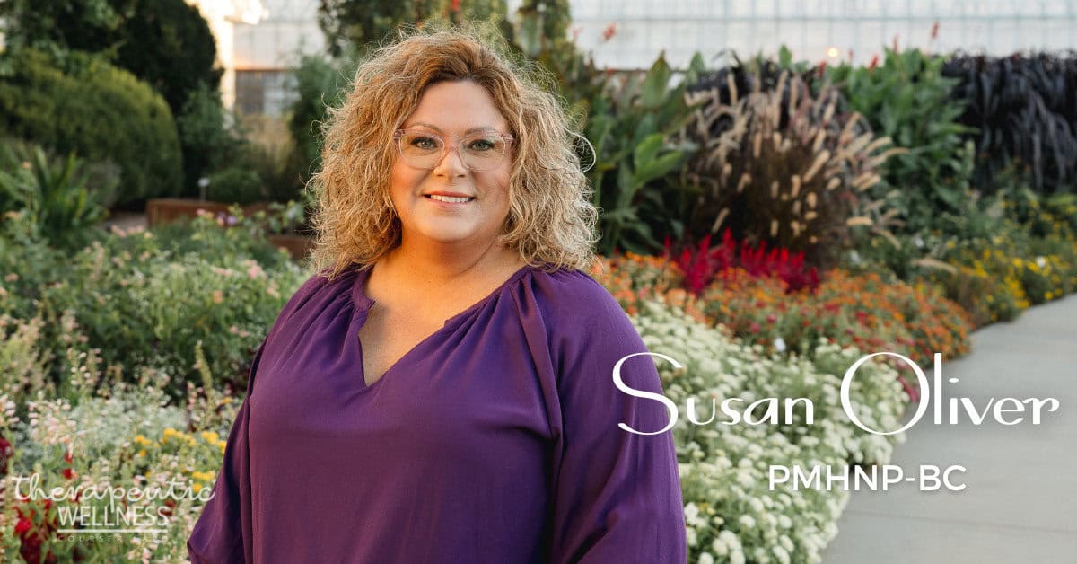 A woman with curly hair and glasses stands smiling in a garden of colorful blooms. Wearing a purple blouse, she exudes warmth. The text on the image reads "Susan Oliver PMHNP-BC" and "Therapeutic Wellness," capturing her compassionate approach to mental health care.