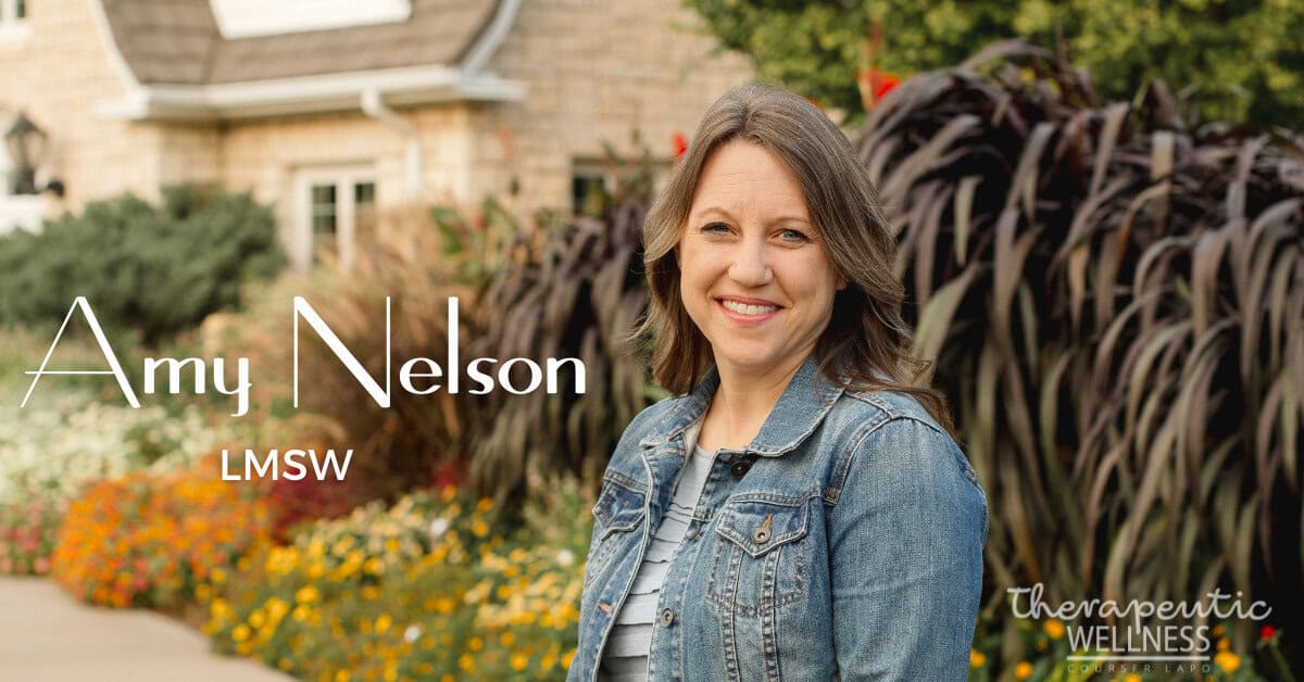 A woman with long brown hair, wearing a denim jacket, smiles while standing in a garden with colorful flowers. Text reads: "Amy Nelson, LMSW. Therapeutic Wellness." A brick house is in the background.