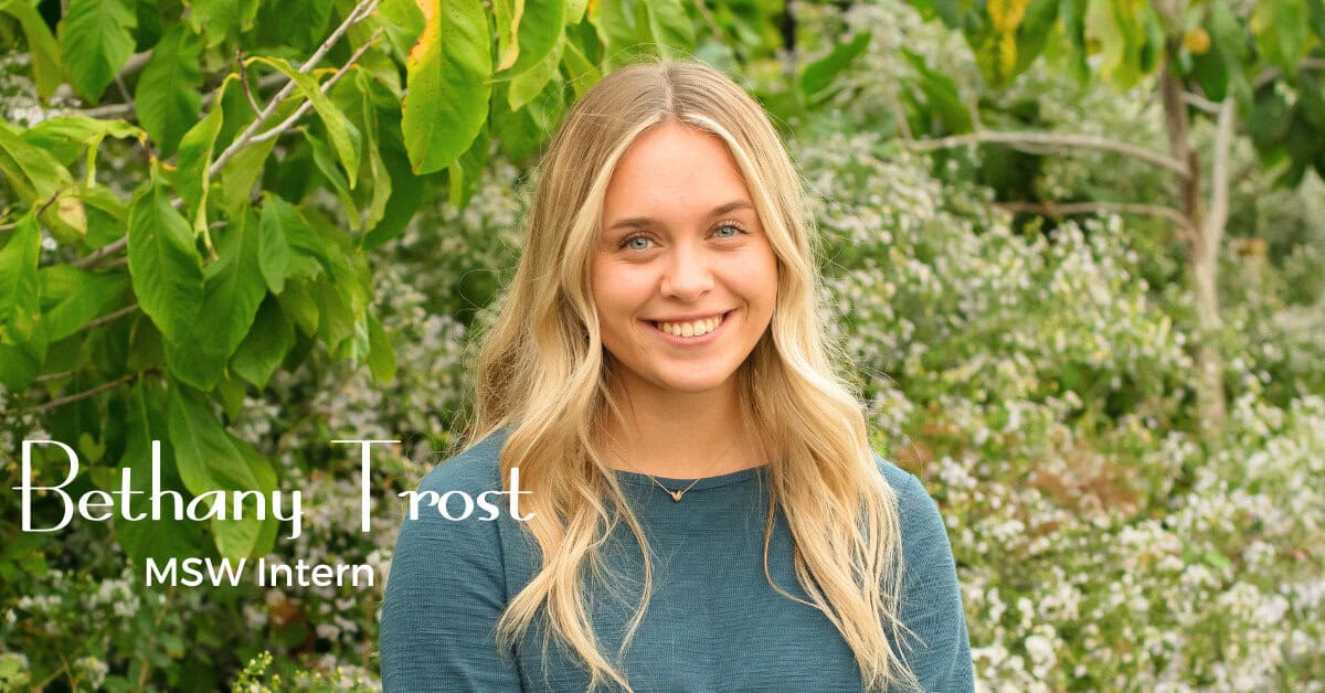 A young woman with long blonde hair wearing a blue top smiles in front of green foliage. Text on the image reads, "Bethany Trost, MSW Intern," introducing Bethany Trost as she radiates warmth and positivity.