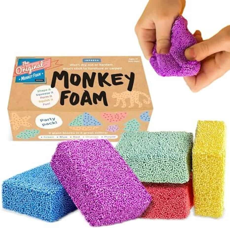 A box labeled "Monkey Foam" with colorful foam blocks&mdash;green, blue, red, yellow, and purple&mdash;stacked in front. A hand squeezes a purple foam block above the box, beside an Expandable Breathing Ball, highlighting sensory fun.