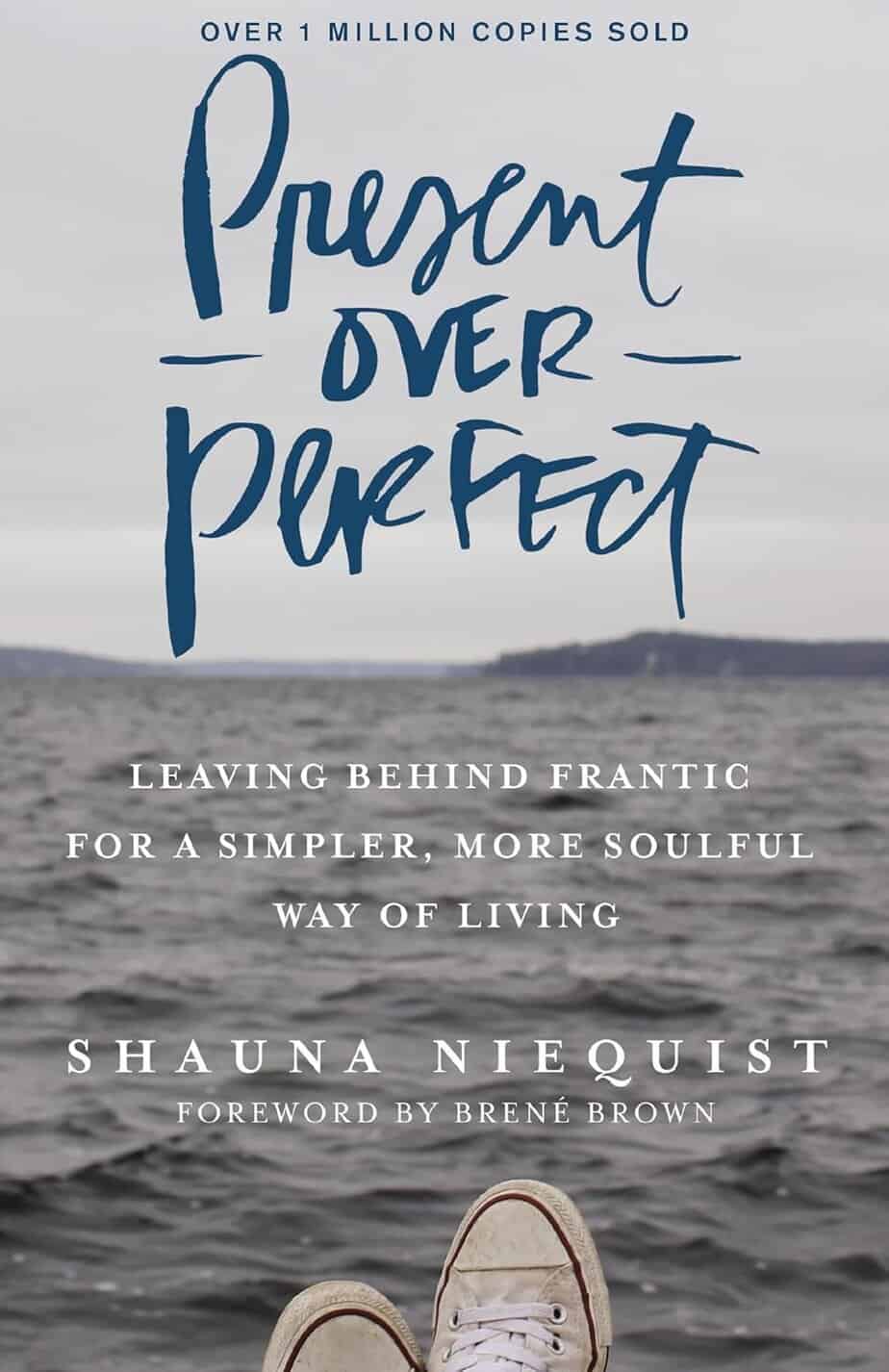 Book cover for "Present Over Perfect" by Shauna Niequist shows a pair of feet in sneakers at the edge of a body of water, with hills in the distance. The subtitle reads, "Leaving behind frantic for a simpler, more soulful way of living.