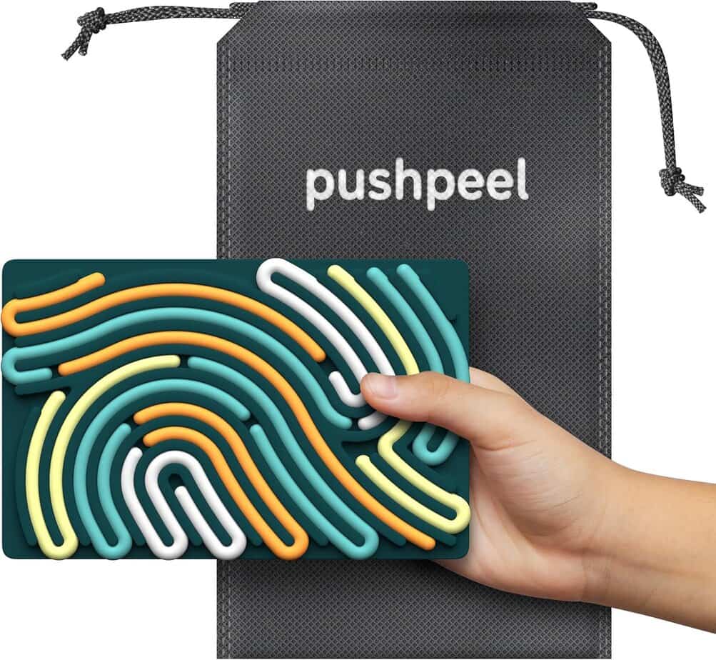 A hand holds a rectangular sensory fidget toy with colorful, raised squiggly lines, perfect for stress relief. Behind it is a gray drawstring pouch labeled "pushpeel" from the playful Grab That Donut collection.