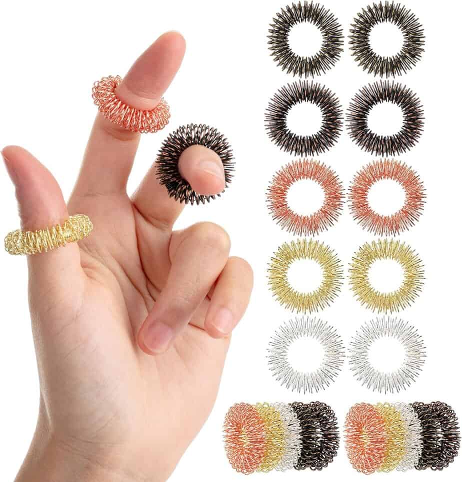 A hand with three spiky acupressure rings on its fingers is shown, alongside a display of multiple rings in black, copper, gold, and silver colors arranged in rows.